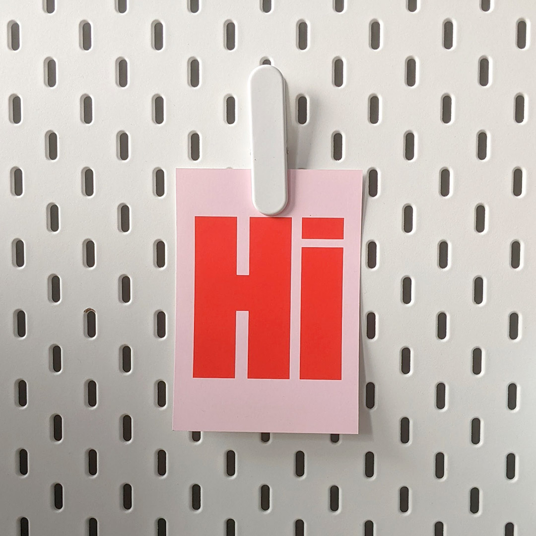 A typographic "hi" postcard in pink and red, clipped to a white pegboard at Yoco Studio, London.